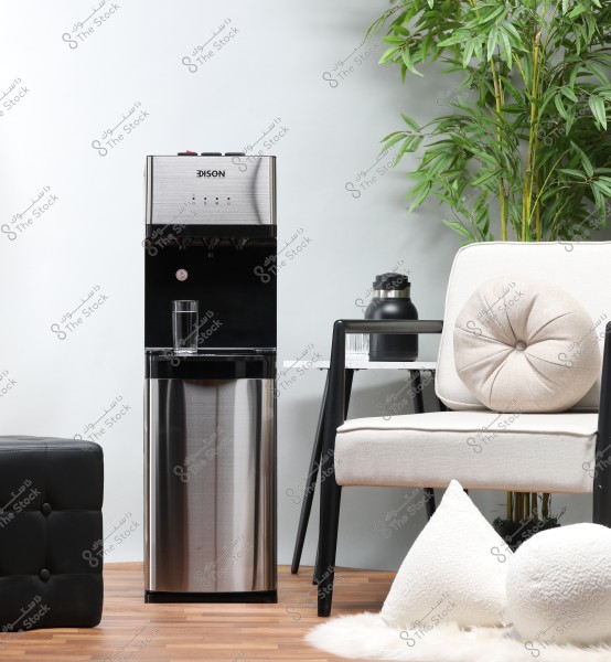 Image showing a silver water dispenser in a modern living room. Surrounding the dispenser are a glass of water, a comfortable beige chair with decorative cushions, and a small table with a thermos against a backdrop of green plants. The wooden floor adds a warm atmosphere.