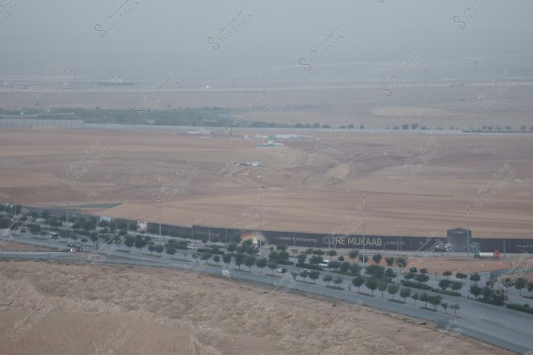 Aerial view of a large construction site in a desert area, showing a wide expanse of land with some construction equipment. A large sign on the perimeter fence displays the slogan \"THE MUKAAB\" with the phrase \"A GATEWAY TO ANOTHER WORLD\". The site is surrounded by several trees along the roadside, and multiple vehicles are seen traveling on the adjacent road.