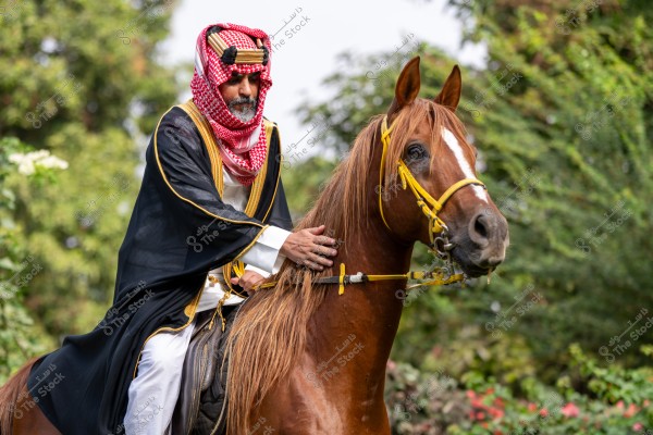 رجل يرتدي الزي التقليدي العربي, بما في ذلك الغترة والعقال والثوب والعباءة السوداء, يركب حصان بني اللون. الخلفية تحتوي على نباتات خضراء وأشجار. الصورة تلتقط جواً ثقافياً وريفياً.
