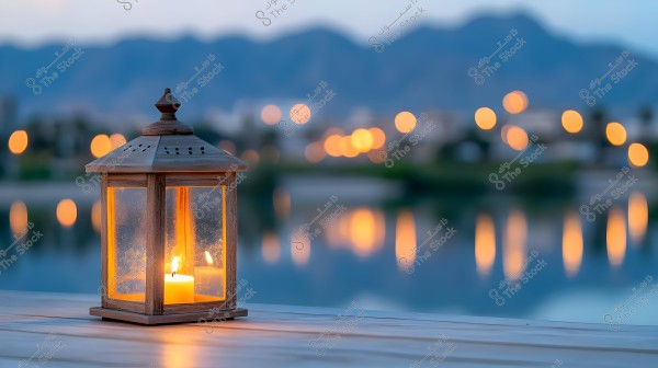 A wooden lantern lit with a candle is in the foreground, placed on a wooden surface next to a lake at sunset. The reflected lights from cities or buildings in the background appear blurred, with distant mountains visible under the dim sky.