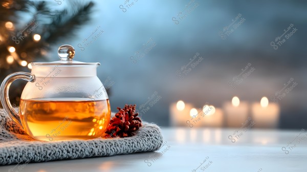 A glass teapot containing a clear golden liquid is placed on a surface covered with a knitted white fabric. Beside the teapot, there is a brown pine cone. In the background, four lit candles are softly blurred on the right, with Christmas tree lights on the left, creating a warm and cozy atmosphere.