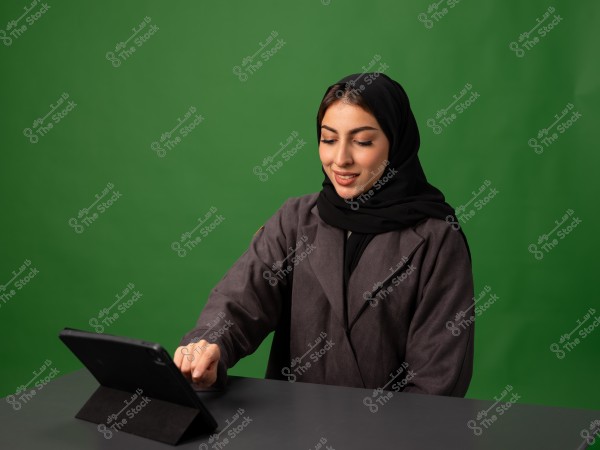 امرأة ترتدي حجابًا وتستخدم جهازًا لوحيًا على طاولة, بخلفية خضراء.