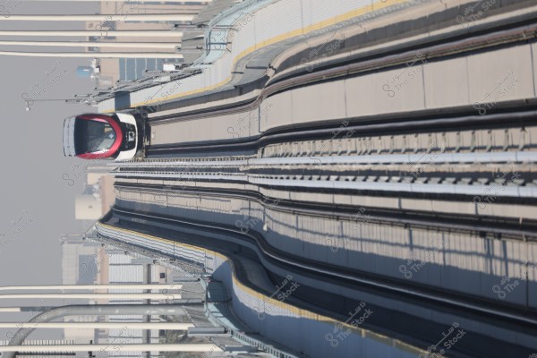 Image of a modern train running on elevated tracks in an urban architectural setting. The train, colored red and black, is moving towards the camera on a dual track. The buildings in the background are blurry, indicating an urban area.