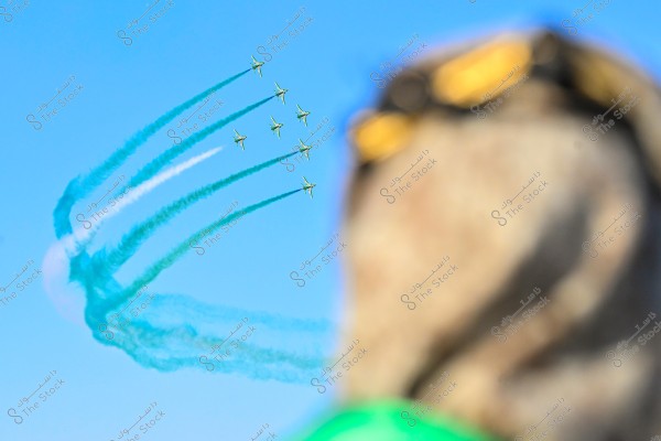 An air show featuring several airplanes emitting sky-colored green and white smoke. In the foreground, there\'s a blurred image of a person wearing sunglasses, a traditional head covering, and green clothing, suggesting a celebratory atmosphere possibly in Saudi Arabia. The sky is clear and blue.