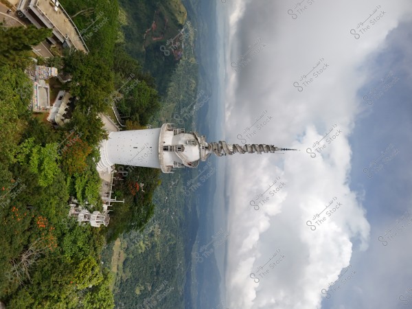 An image of a tall tower with a spiral top, surrounded by lush greenery and dense trees. The background features a blue sky with white clouds, adding a serene and natural touch to the scene.