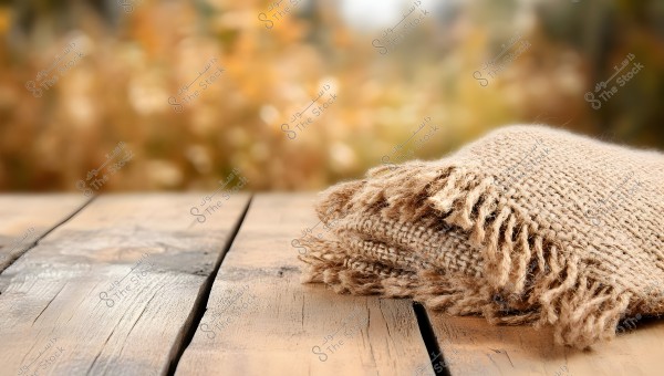 An image featuring a light brown woolen scarf placed on a wooden table outdoors. The fringed edges of the scarf add a cozy, rustic touch to the scene. The background is a blurry autumn setting with golden and brown colors, creating a warm and serene atmosphere.