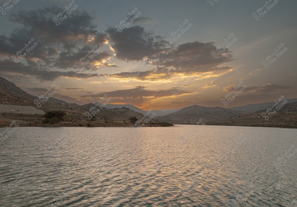 A natural landscape featuring a lake surrounded by hills in the distance, with a sky filled with scattered clouds. The sunset casts shades of orange and yellow across the sky. Reflections on the lake\'s water enhance the beauty of the scene.