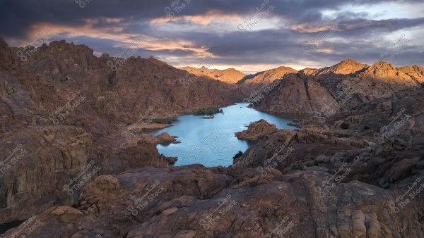The image depicts a natural landscape of a lake surrounded by massive rocky mountain formations under a cloudy sky. The rocks are in warm earthy tones, while the water reflects the grayish-blue color of the sky. The horizon of the mountains is bathed in the setting sun\'s rays, adding a golden glow to the scene.