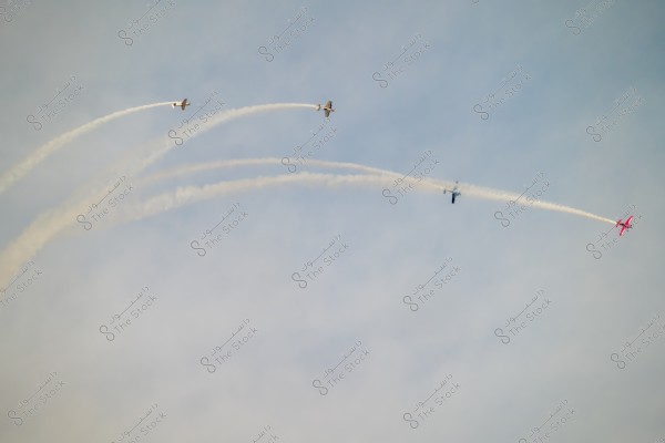 The image shows four airplanes flying in the sky, leaving smoke trails behind them. The airplane at the front is painted pink, while the others are in different colors. The white smoke curls across the clear sky in an aerobatic display.