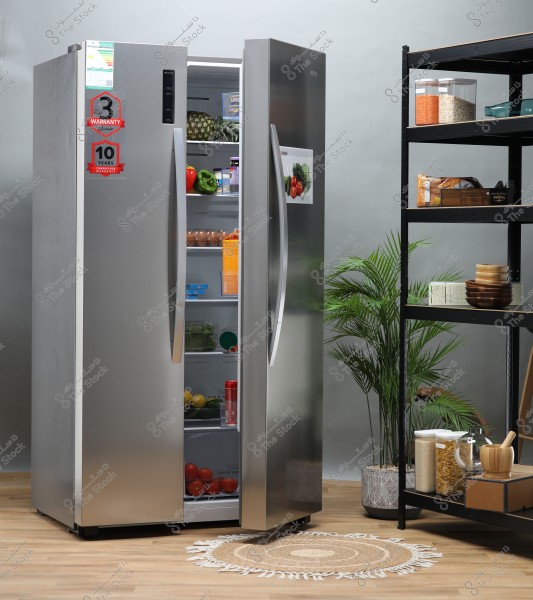 Image of a modern kitchen featuring a stainless steel refrigerator partially open, containing fruits and vegetables like pineapple, tomatoes, bell peppers, and lettuce, along with juice cartons and eggs. Next to it is a metal shelf holding containers of legumes and some kitchen utensils. There is also a decorative green plant on the wooden floor, and a small decorative rug next to the refrigerator.