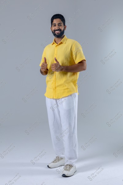 An image of a man standing and smiling, wearing a yellow shirt, white pants, and white sneakers. He appears to be giving thumbs up, representing a positive and cheerful demeanor. The background is simple and light gray in color.