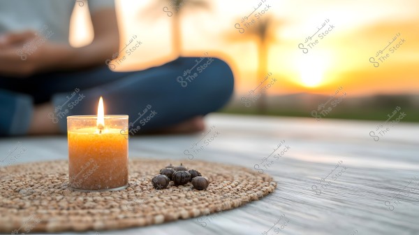 An image featuring a lit candle on a woven mat alongside some black seeds. In the background, a person is seated on the ground wearing jeans and a white shirt. The backdrop displays a sunset with blurred palm trees.