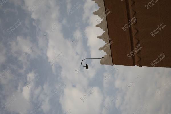 Image of a section of a traditional building wall with white triangular motifs at the top and a simple metal lamp hanging from the summit. Part of the building appears to be made of mud or traditional materials in a light brown color, with a background of a sky filled with white clouds.