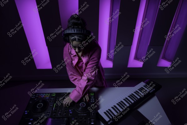 دي جي ترتدي معطفًا ورديًا تعمل على طاولة خلاط بجانب لوحة مفاتيح إلكترونية تحت إضاءة بنفسجية.