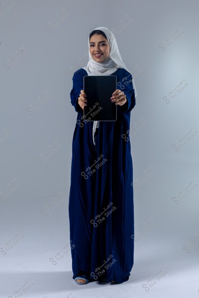 An image of a woman wearing a blue abaya and a white hijab, holding a tablet towards the camera with a grey background. She appears to be from the Gulf region.