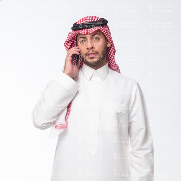 بورتريه رجل سعودي يرتدي الثوب ويقف أمام خلفية بيضاء ويتحدث عبر الجوال