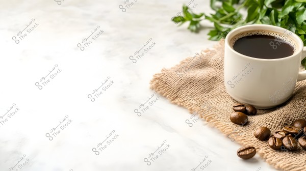 A white coffee cup filled with black coffee placed on a burlap cloth on a white surface. Next to the cup are coffee beans and some green leaves in the background.