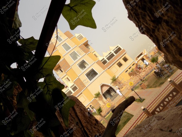 Image of a multi-storey traditional building prominently displaying architectural design inspired by Arabian heritage, characterized by beige colors and geometric black and white patterns along its edges. Green plant leaves appear in the foreground, while people dressed in traditional white clothing are present in the courtyard in front of the building. The weather appears sunny with a clear sky.