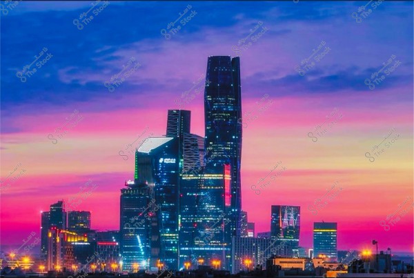 Image of an urban scene in Riyadh at sunset, showing the city skyline with skyscrapers illuminated with yellow and white lights. The sky transitions from blue to pink and purple, creating a stunning backdrop behind the modern buildings.