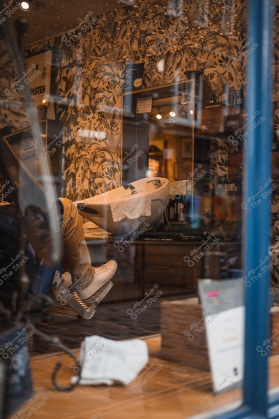 View through a barber shop window showing the feet of a person sitting on a barber chair inside. The interior decor features floral patterned wallpaper.