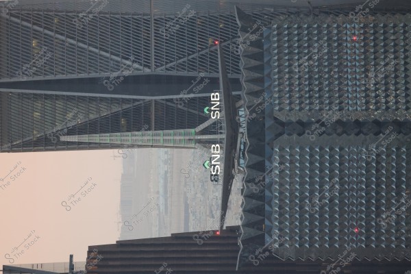 An image of tall skyscrapers in the city, featuring a modern angular architectural building with closely aligned and symmetrical windows. The building has signage with \"SNB.\" It showcases innovative geometric designs with glass materials and sharp angles. The background sky appears in a dull gray color, suggesting dusk or an overcast day.