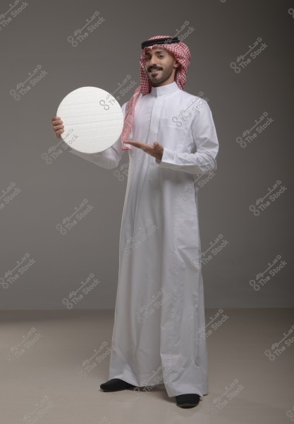 A portrait of a man wearing a white thobe, a red checkered headscarf, and a black agal. He is smiling and pointing to a white circle he holds in his hand. The background is light gray.