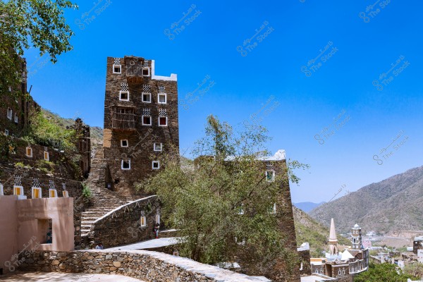 The image shows multi-story traditional stone houses located in a mountainous area. The buildings are made of stone and have square windows with white and black patterns. A large green tree is prominently in front of the buildings, and the mountainous landscape is visible in the background under a clear blue sky.