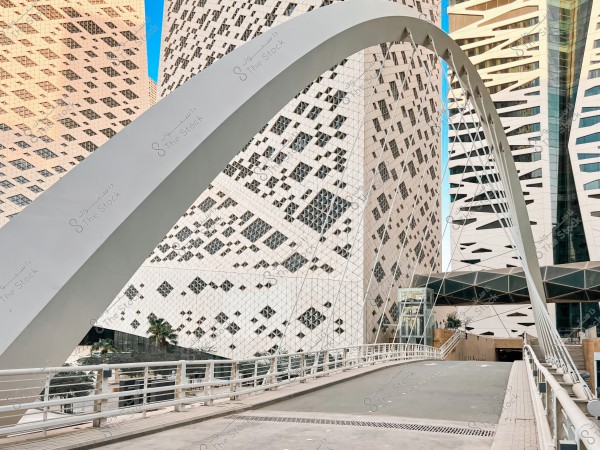A modern bridge with white arches extends towards a cluster of buildings with intricate architectural designs and geometric patterns. The sky is blue, with palm trees visible below between the buildings.