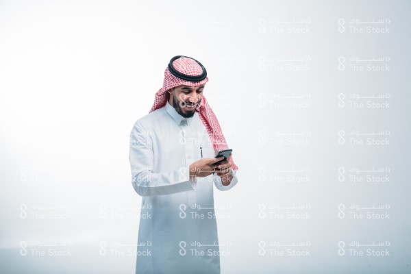 رجل سعودي يرتدي الزي الرسمي السعودي على خلفية بيضاء ، يمسك الجوال ، يستخدم أحد تطبيقات الآيفون ، يتصفح الإنترنت ، يستخدم وسائل التواصل الاجتماعي