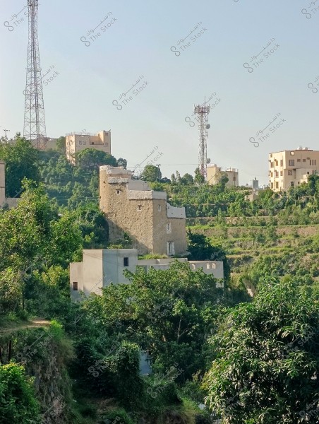 منظر طبيعي من محافظة فيفاء بمنطقة جازان جنوب المملكة العربية السعودية لمباني تقع في منطقة جبلية خضراء. تظهر في الصورة منازل تقليدية مصنوعة من الطوب, وأبراج اتصالات تمتد في الخلفية. الأشجار والنباتات الخضراء تحيط بالمنازل, مما يشير إلى بيئة طبيعية وجبلية. يوجد مبنى تقليدي يشبه الحصن بالقرب من مقدمة الصورة.