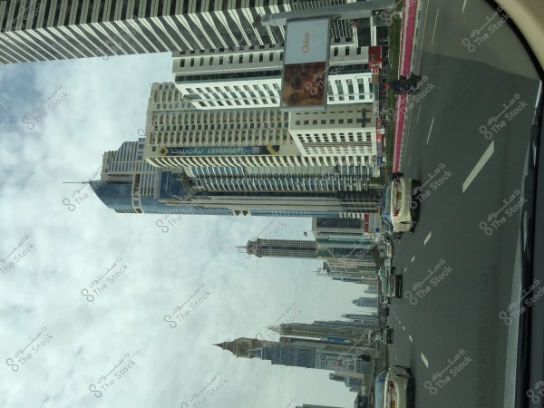 An image depicting a major street in a modern and upscale city, surrounded by various skyscrapers with modern glass architectural designs. There is an advertising billboard on the left side, and flags are visible atop some of the towers. Cars are shown driving on the multi-lane road, and the sky is covered with clouds.