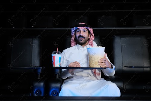 شاب سعودي يرتدي الزي السعودي التقليدي  يجلس في صالة السينما ويتفاعل أثناء مشاهدة الفيلم، رجل سعودي يرتدي الزي السعودي التقليدي يشاهد أحد الأفلام المعروضة في صالة السينما بحماس ويظهر أمامه الفشار وأحد المشروبات على الطاولة