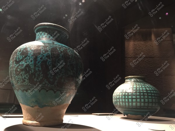 The image shows a large vase decorated with Arabic calligraphy in blue and teal colors, alongside a smaller vessel with a grid pattern in similar colors, placed on a display table in a dimly lit setting.
