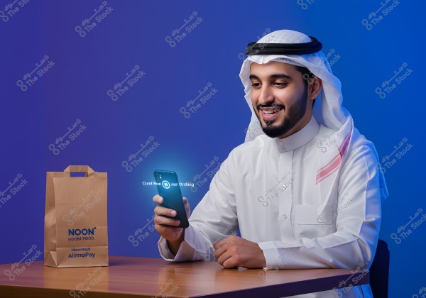 The image shows a man wearing a white thobe and a ghutra with an iqal, sitting at a table. The photo appears to be taken in a studio with a blue-lit background. The man is looking at a smartphone in his hand with a smile, and there is a brown bag in front of him with the \"Noon Food\" logo.