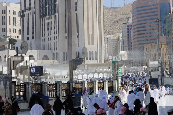 A group of pilgrims wearing white Ihram garments walking in an outdoor courtyard near the Grand Mosque in Mecca, Saudi Arabia. In the background, several high-rise buildings surround the area, alongside a sign indicating a section for people with disabilities. There are many other people in various attire, including women in hijabs, and the scene is sunny with cooling devices visible.