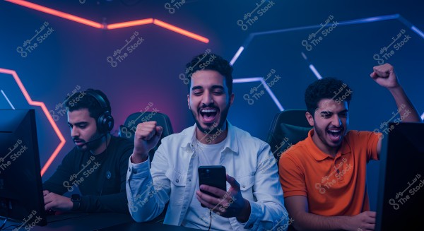 The image shows three young men in a modern gaming room. The first young man on the left is wearing a headset and sitting in front of a computer screen, appearing focused on the game. The middle young man is wearing a white shirt and holding a smartphone, expressing great joy with a raised fist and a wide smile. The young man on the right is wearing an orange shirt and also celebrating with a raised fist. The background features blue and red neon light lines.