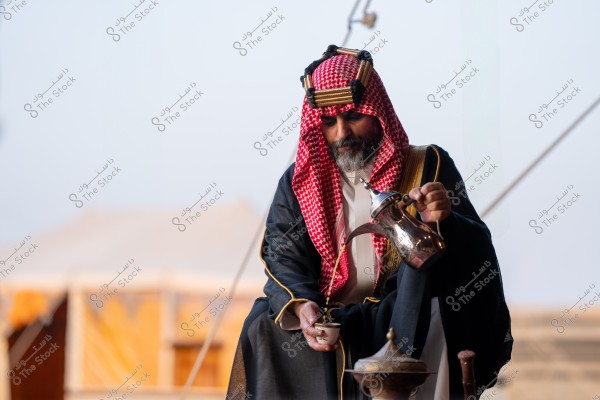 صورة لرجل جالس يرتدي الثوب العربي التقليدي مع الشماغ الأحمر والعقال, ويصب القهوة من الدلة العربية في فنجان صغير. تظهر خلفه بعض تفاصيل الخيمة التقليدية في الهواء الطلق.