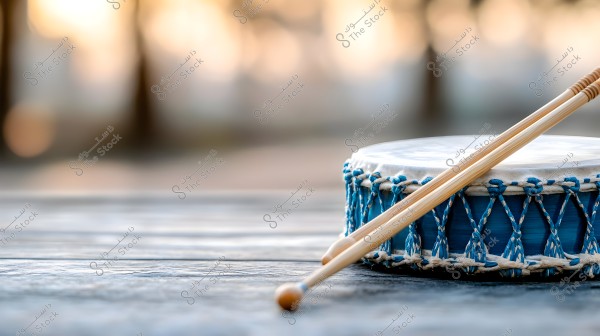 An image of a small blue drum placed on a wooden surface. The drum is decorated with blue and white strings and patterns, with two wooden sticks resting on it. The background is blurred with light colors, highlighting the details of the drum.