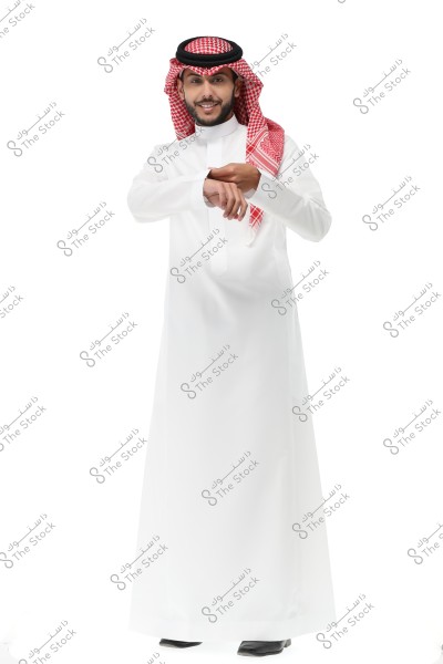 A man wearing a white thobe and a red-checkered ghutra with a black agal. He is smiling and looking at the camera, with his hands placed on a wristwatch. The thobe and ghutra are part of traditional Gulf attire, commonly worn in Saudi Arabia.