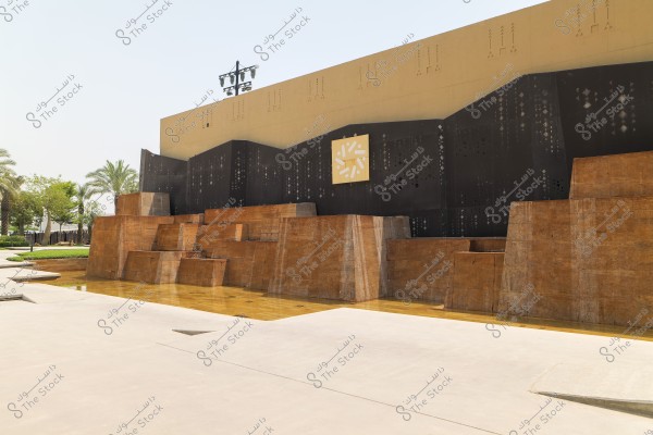 The image shows a brown architectural wall with multiple layers and geometric details. Behind the wall is a modern beige building with black decorative designs, and at the top, there is a yellow panel featuring a white decorative pattern. The image also includes palm trees and greenery in the background.