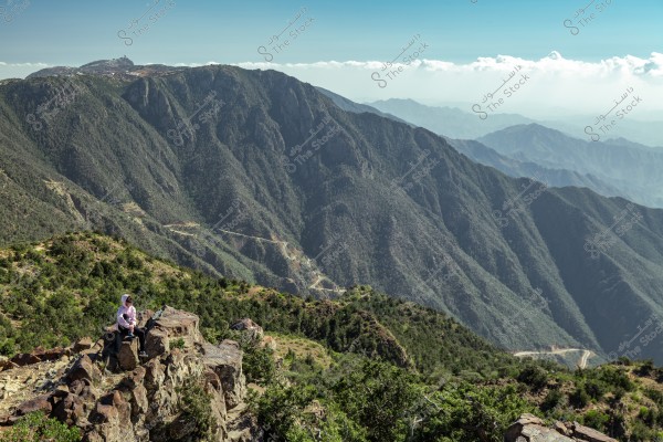 A daytime landscape showing a high mountain covered in green trees. A winding road can be seen weaving through the mountains. In the foreground, a person is sitting on rocks, wearing traditional Saudi clothing and a white headscarf, enjoying the expansive panoramic view.