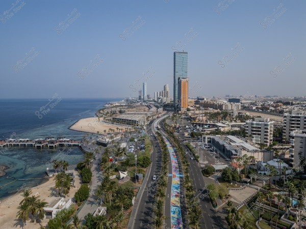 Aerial view of Jeddah, Saudi Arabia, showcasing the coastal road and corniche along the Red Sea. The image features tall buildings, a sandy beach, and green areas. The road is adorned with artistic decorations on one side, with trees, streets, and urban developments interspersed throughout.