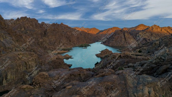 A natural landscape showing rugged mountainous terrain with a lake in the center. The clear water is surrounded by reddish-brown rocky hills. The sky is blue with scattered white clouds.