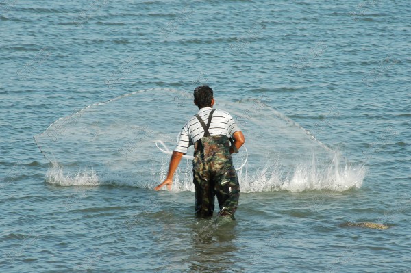 A man wearing green and gray fishing attire stands in shallow water, casting a circular fishing net into the sea. The water ripples around the net as it\'s thrown. The background consists of an expanse of blue water.
