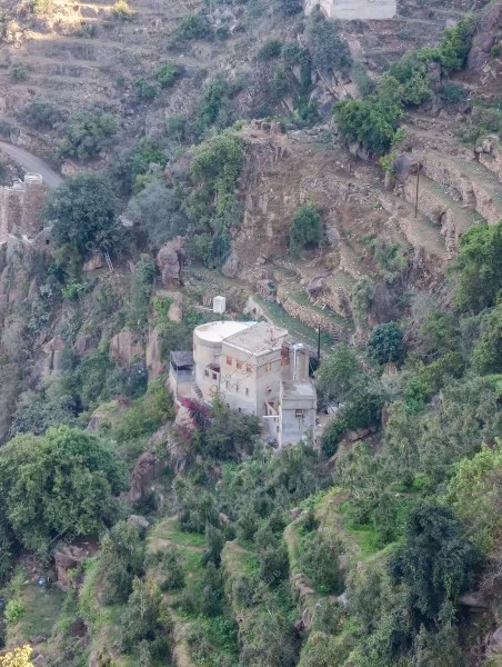 منزل تقليدي مبني من الطوب والإسمنت يقع في منطقة جبلية خضراء. تحيط به الأشجار الكثيفة والمدرجات الزراعية. تظهر المرتفعات الصخرية في الخلفية مع طريق ضيق يؤدي إلى المنزل.