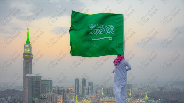 A snapshot of a Saudi man wearing the traditional Saudi dress, carrying the Saudi flag over a mountain in the Makkah Al-Mukarramah region, and the sky appears clear, the royal clock tower building in the Grand Mosque, buildings and landmarks, the Grand Mosque of Mecca.