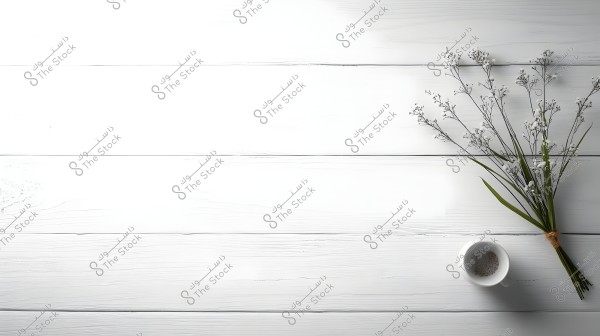An image of a white wooden table surface featuring a partially empty white cup with residue powder inside, alongside a flowering branch with small white flowers bundled together with a simple twine.