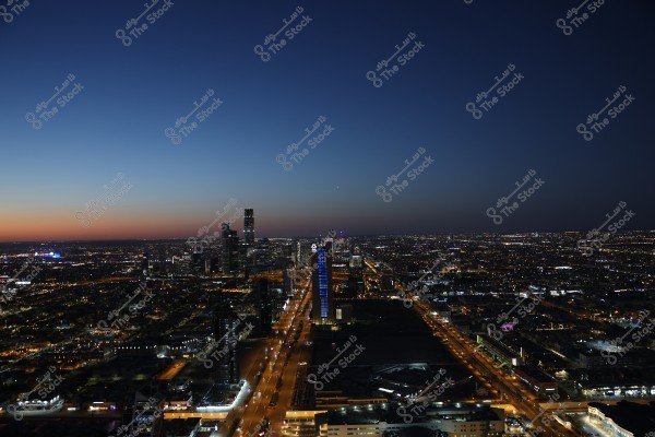 Aerial view of a city at night with a lit skyline under a dark sky fading to a lighter hue at the horizon. The image shows illuminated skyscrapers and long roads lit by streetlights. The scene likely depicts a modern city in the desert, such as Riyadh in Saudi Arabia.