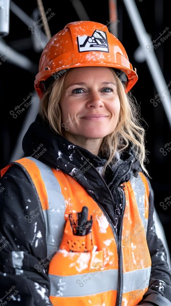 صورة بورتريه لامرأة تبتسم, ترتدي سترة أمان برتقالية اللون وخوذة برتقالية في موقع بناء. السترة تحتوي على طلاء أبيض وعلامات أمان. في الخلفية, يظهر هيكل غير واضح.