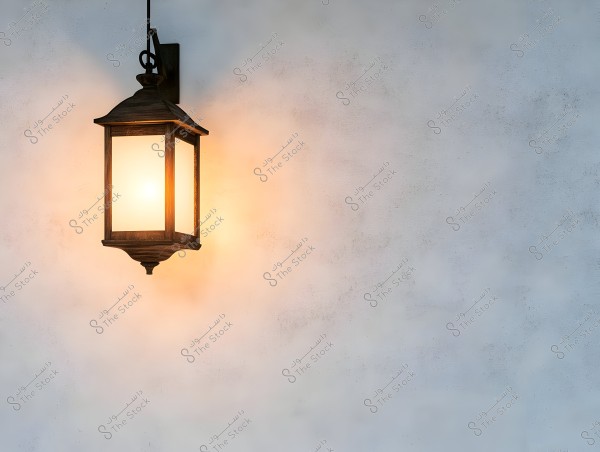 An antique metal lantern hangs on a light-colored wall. The lantern is rectangular and emits a warm light that glows around it. The surrounding wall is smooth and appears in soft hues ranging from light blue to gray.